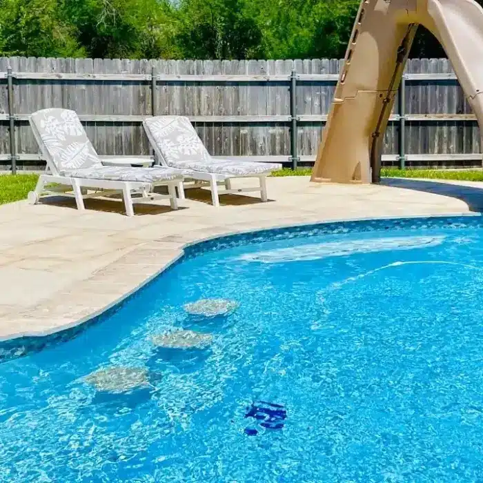 A backyard swimming pool with two lounge chairs, a slide, and a wooden fence in the background.