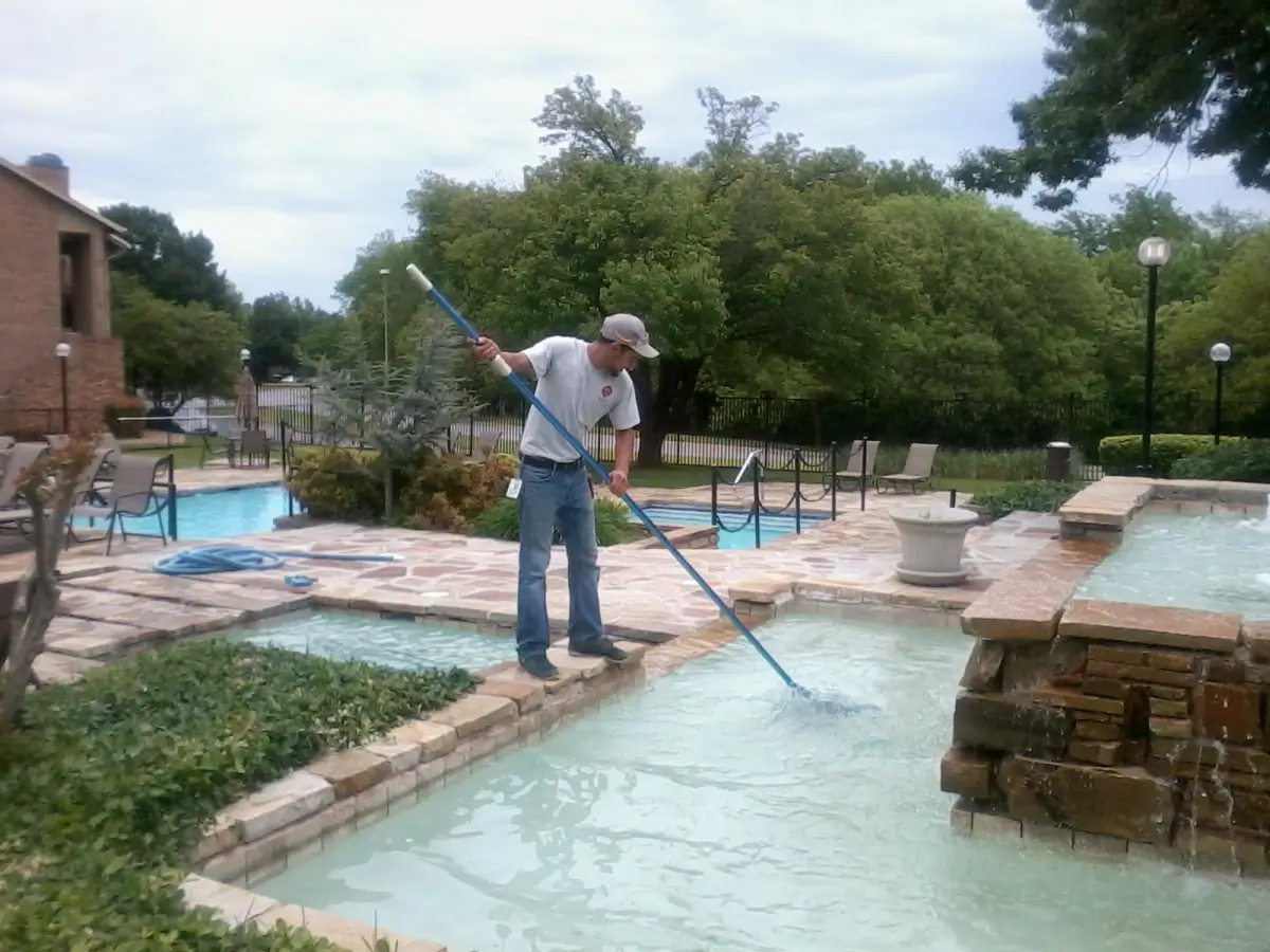 Man cleaning a swimming pool with a long-handled brush, surrounded by garden and patio areas.