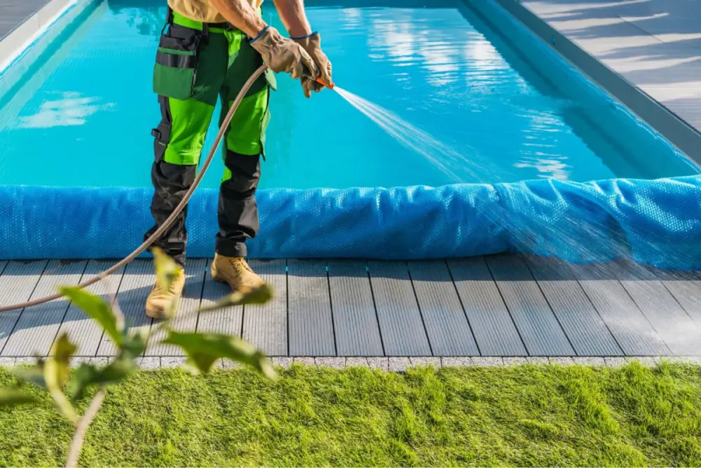 Worker wearing gloves and protective gear using a pressure washer near a swimming pool.