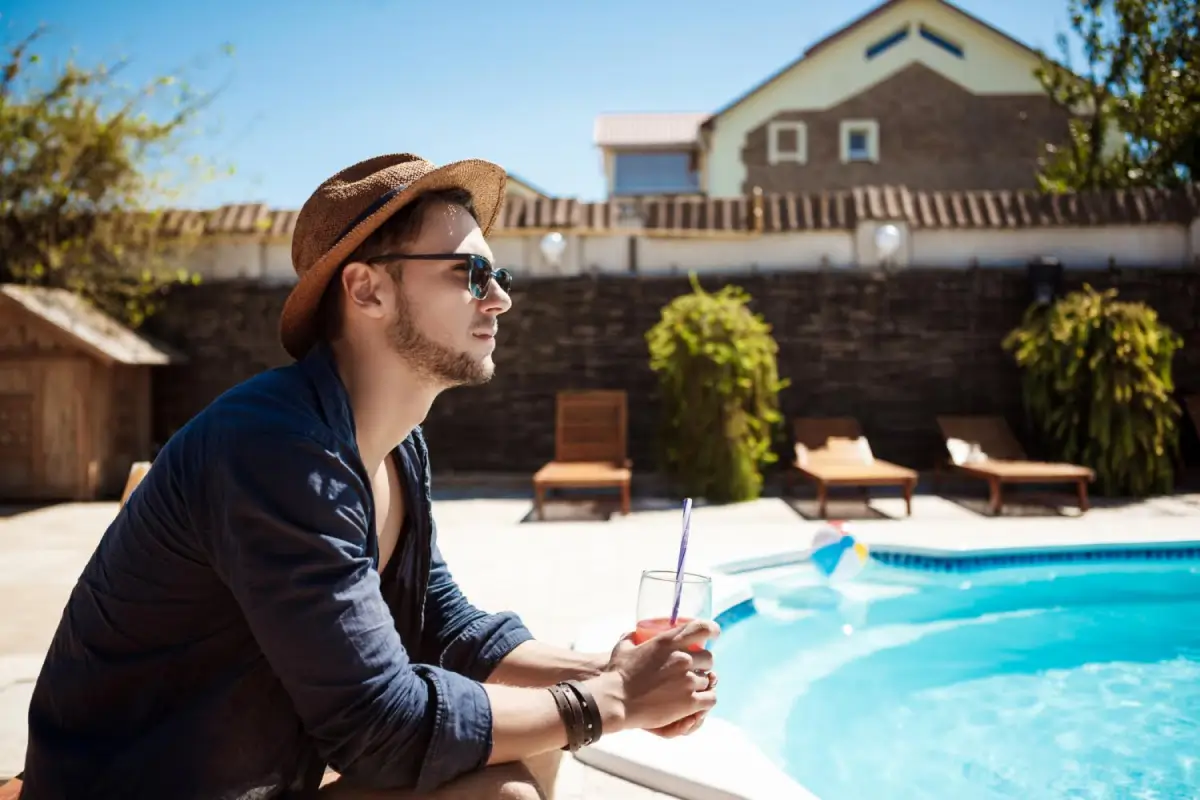 Man relaxing by the poolside, wearing a hat and sunglasses, holding a drink.