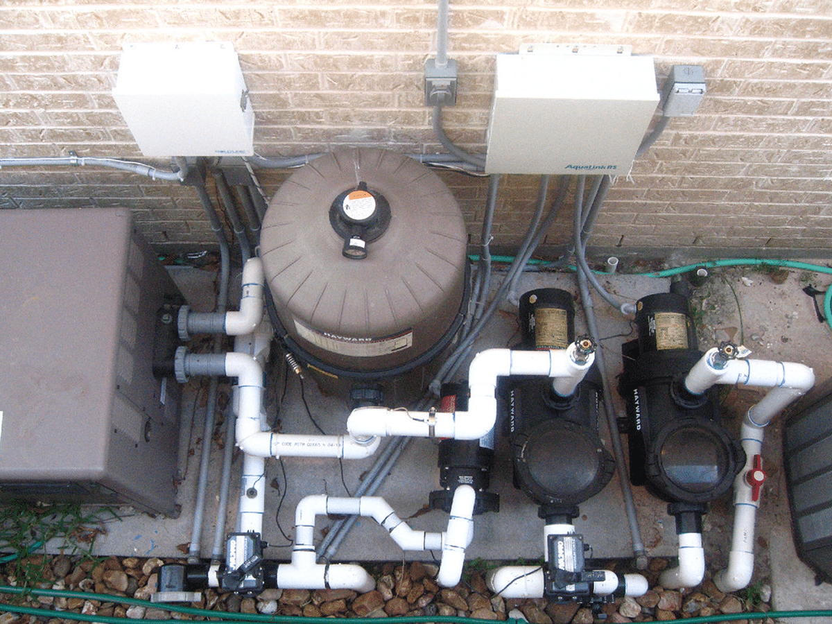 A complex outdoor pool filtration system with multiple pipes and equipment attached to a brick wall.