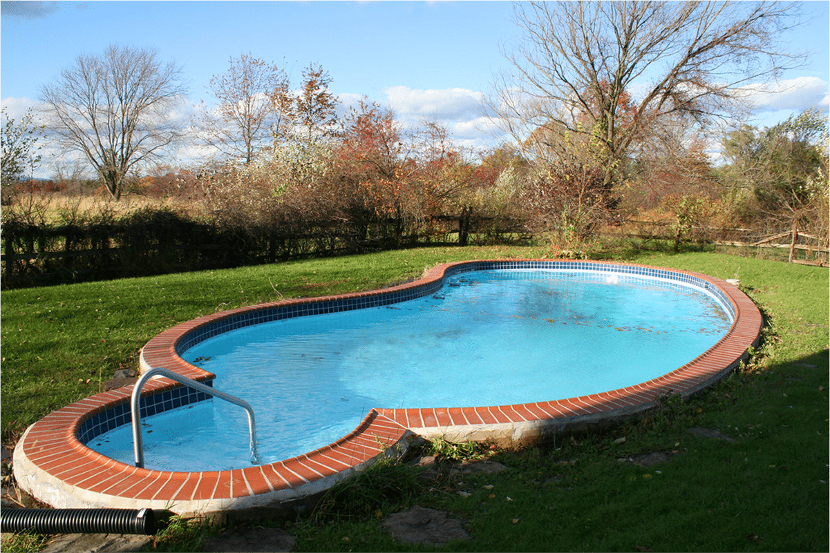 A kidney-shaped pool with red brick trim, surrounded by grass and trees.